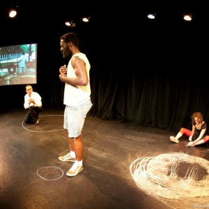 Ali and Tiphaine on floor beside chalk drawn circles. Pete stands in middle behind small chalk circle; projected photo on back wall - image of playground with tire swings.